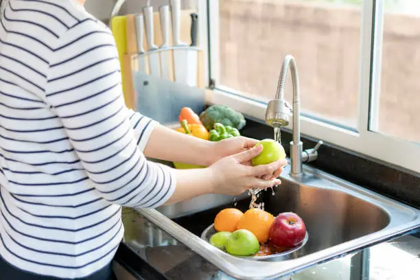 Photo of Asian healthy woman washing an apple and other fruit above kitchen sink and cleaning a fruit / vegetable with water to eliminate the chances of contamination COVID-19.