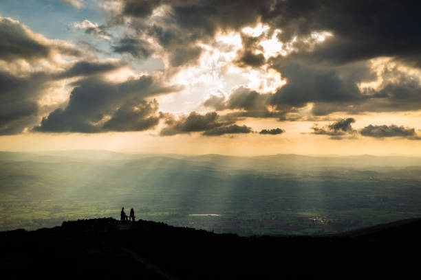 A couple sit on a bench during dramatic sunset over countryside with hill and mountain stock photo