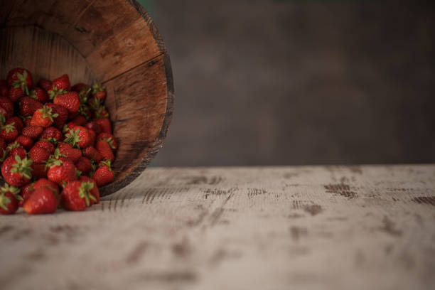 Cut out of the wooden rustic bucket placed sideways on top of the kitchen table to show the load of beautiful, red strawberries falling outside and on the table.