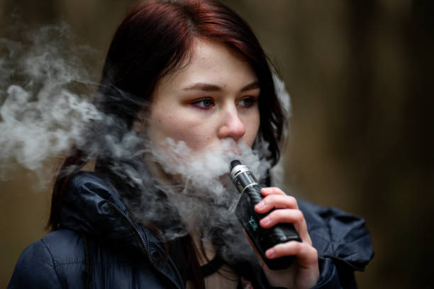 adolescente vapeo. joven chica morena bastante caucásica fumando un cigarrillo electrónico en la calle en la primavera. mal hábito. - cigarrillo electrónico fotografías e imágenes de stock