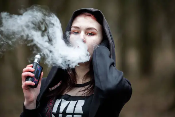 Vape teenager. Young pretty caucasian brunette girl with problem skin in the hood smoking an electronic cigarette on the street in the spring. Deadly bad habit.