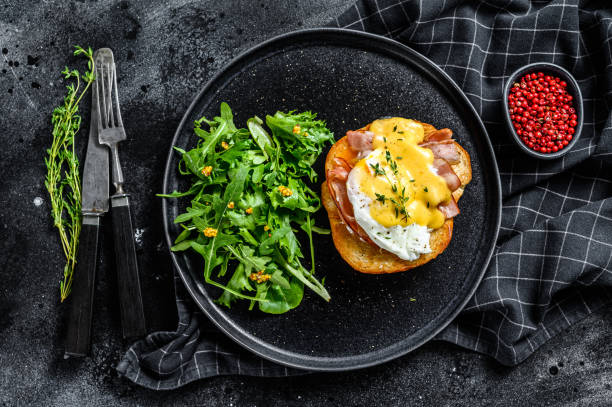 istock Brioche sandwich with bacon, egg Benedict and hollandaise sauce. Garnish with arugula salad. Black background. Top view 1221984176