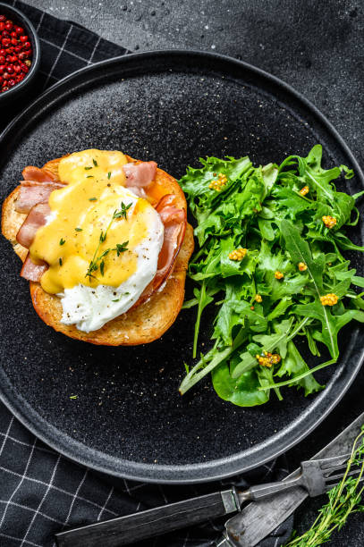 istock Brioche sandwich with bacon, egg Benedict and hollandaise sauce. Garnish with arugula salad. Black background. Top view 1221984374