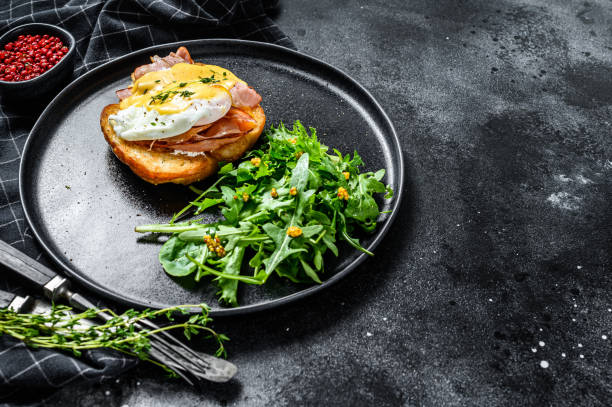 istock Brioche sandwich with bacon, egg Benedict and hollandaise sauce. Garnish with arugula salad. Black background. Top view. Copy space 1221984437