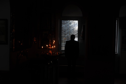 Silhouette of a priest standing before a bright window in the dark room. Candles in old medieval christian church. Religion, christianity and belief concept.
