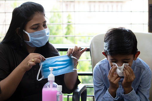 Madre cautelosa sosteniendo máscara médica para el hijo estornudando en casa photo