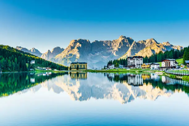 View of a small town surrounded by a pine forest through a mountain lake. Massive Italian Alps on the background.