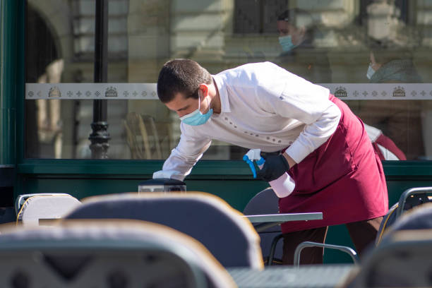 camarero con una máscara desinfecta la mesa de un bar, cafetería o restaurante al aire libre - leaving business landscape men fotografías e imágenes de stock