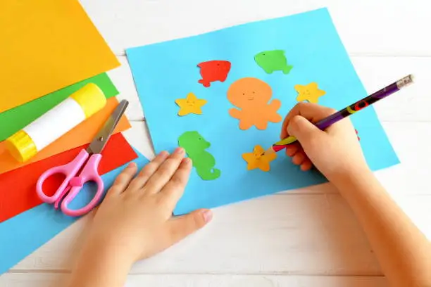 Photo of A child holds a pencil and draws. The child doing a card with sea creatures and fish. Sheets of colored paper, scissors, glue, set for easy paper summer crafts for kids