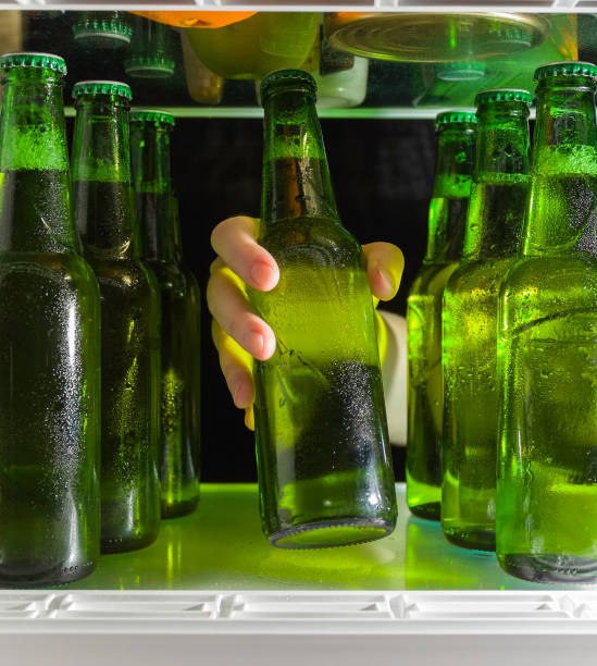 la mano raggiunge la bottiglia di birra. bottiglie verdi di birra e gocce di condensa sono su uno scaffale in frigorifero. - beer bottle beer cold alcohol foto e immagini stock