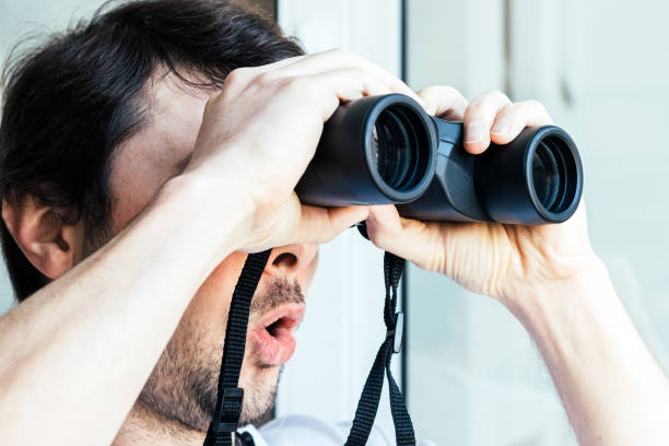 istock Handsome man with a surprised face looking through binoculars at the window with a view of the neighboring house for observing neighbors, environment or nature. Selective focus. Closeup view 1222001305