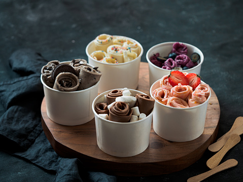 Rolled ice cream in cone cups on rustic round wooden tray. Different thai style rolled ice cream on datk background