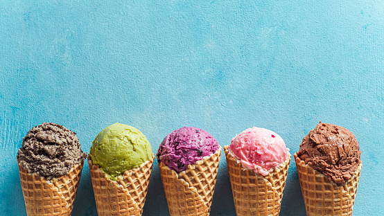 Various ice cream scoops in cones with copy space. Colorful ice cream in cones chocolate, strawberry, blueberry, pistachio or matcha, biscuits chocolate sandwich cookies on blue background. Top view