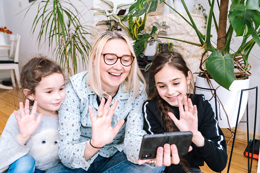Woman with children sitting at home and holding videocall. Family using smartphone for video call with friend or family. People communicates via video - looking camera and waving greeting hands.