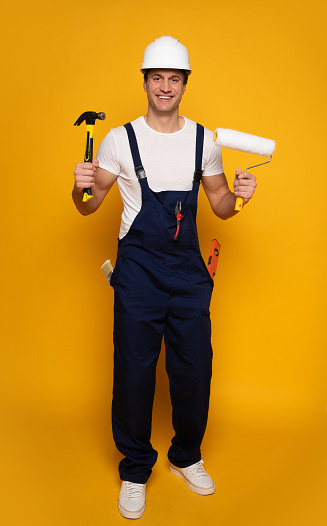 Carpenter. Full-length photo of a man in a blue working suit and  helmet, who is looking in the camera while holding a hammer and a painting roller.