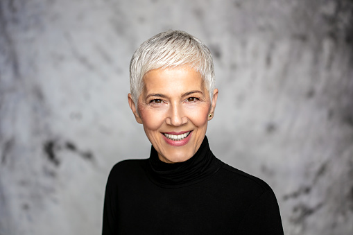 Close up portrait of beautiful mature woman with grey hair wearing black turtleneck looking at camera and smiling. Attractive middle aged woman with beautiful smile isolated over grey background.