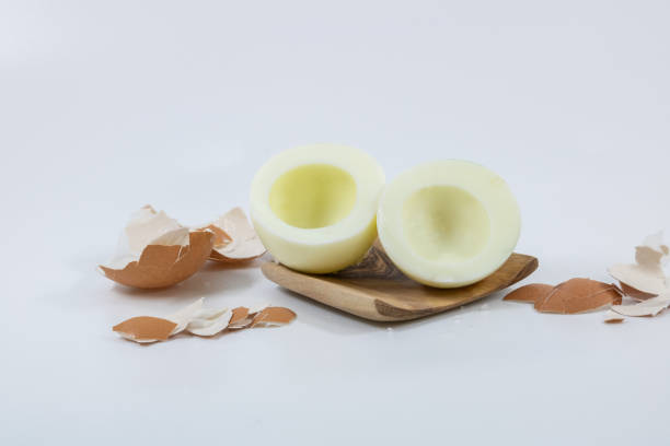Hard-boiled egg white hollowed out of its yolk in a wooden spoon with egg shell around, photo on white background with negative space stock photo