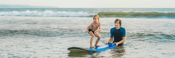 Father or instructor teaching his 4 year old son how to surf in the sea on vacation or holiday. Travel and sports with children concept. Surfing lesson for kids. BANNER, LONG FORMAT