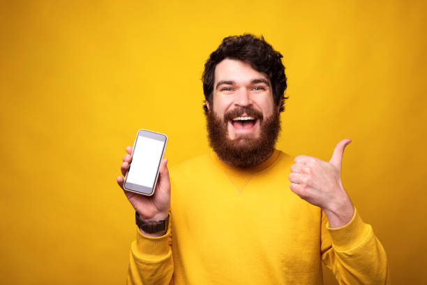 Happy man shows thumb up or like gesture while holding his phone with blank screen on yellow background.