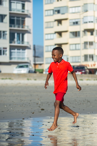 A young African Boy running barefoot on beach and wet sand side view.  He is wearing a bright red shorts and golf shirt.