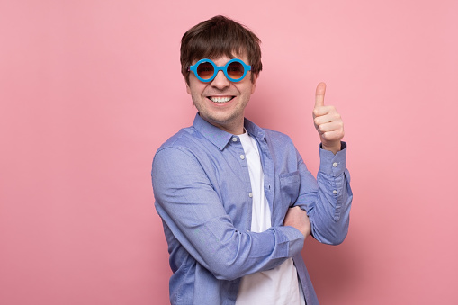 Happy caucasian man in summer glasses with thumbs up gesture. Positive facial human emotion, studio shot on pink wall