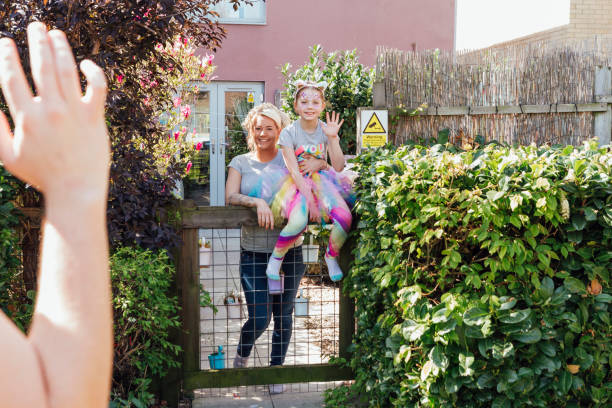 Waving To The Neighbor A young Caucasian girl wearing a tutu and rainbow colors, sits on top of the garden gate with her mother standing beside her, they wave to their neighbor at a safe distance. neighbour stock pictures, royalty-free photos & images