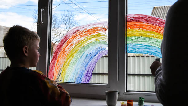 istock Little boy painting rainbow on a window and looking through the window while sitting home during quarantine. 1222042468