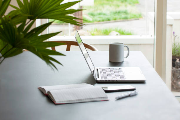 istock Laptop computer on a table with coffee mug, notepad, mobile phone and pencil. A green palm flower in the foreground and view outside a window to a garden. Working or studying at home concept. 1222052879