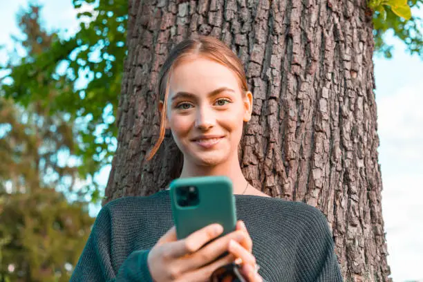 Happy smiling young woman leaning on tree in the garden, reading messages on her mobile phone, looking up, smiling happy towards the camera. Young women social media lifestyle concept shot.