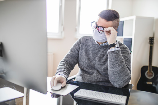 Young businessman having headache and wearing protective face-mask and surgical glove at office