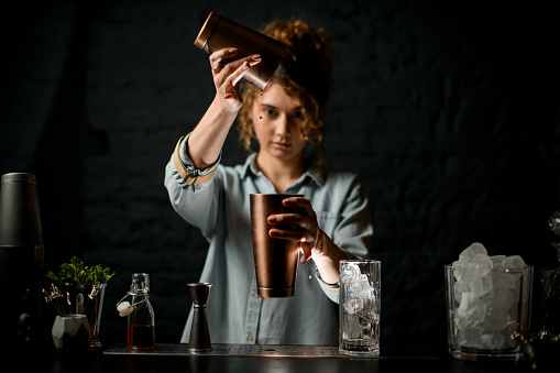 young girl at bar pours drink from one metal cup to another and looks at it. Different bar equipment stands nearby.