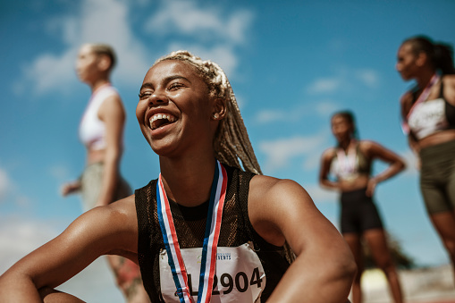 Female athlete smiling after winning a race with other competitors in background. Sportswoman with medal celebrating her victory at stadium.