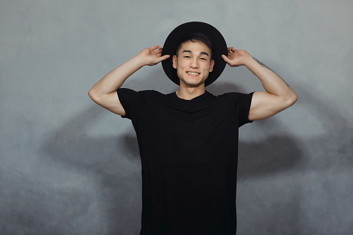 Handsome Asian man with short haircut posing in a trendy black hat and black t-shirt against gray background in the studio. Indoor photo of a confident guy in stylish black clothing looking at camera