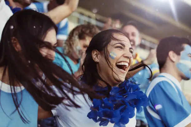 Crowd of sports fans of Argentina cheering during a match in stadium. Group of friends watching sports game celebrating when their team scoring a goal.