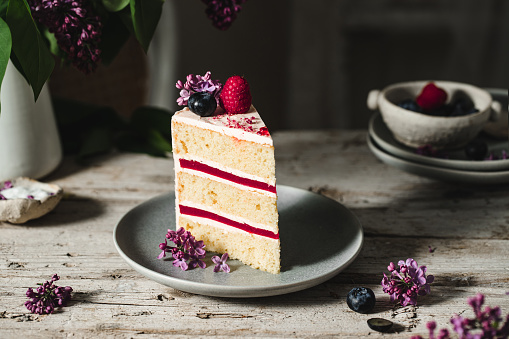Slice of a homemade cake decorated with berry fruits and lilac flowers. Delicious cake served on a table.