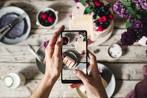 POV shot of a female chef hands taking photograph of a freshly make cake with her mobile phone. Woman taking pictures of a fruit and flower cake served on table.