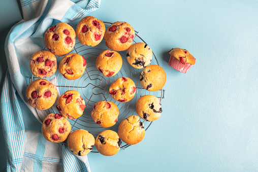 Oven fresh muffins with berries, on a cooling rack, on a blue table. Above view of mixed muffins with blueberry, raspberry, and strawberries.