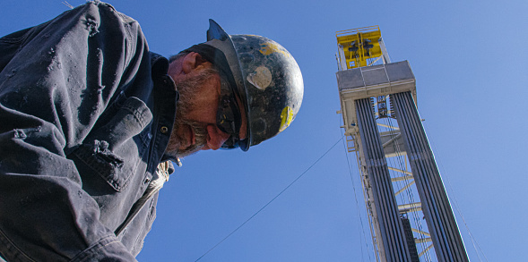 A Male Oilfield Worker in His Sixties Works Next to a Derrick at an Oil and Gas Drilling Pad Site on a Sunny Morning