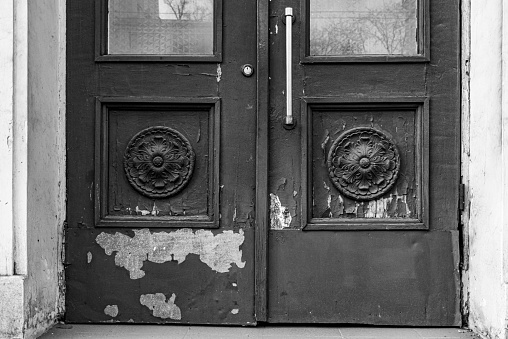 Black and white photo of architectural fretwork elements of old door with flaking paint and stains. Sculptural rosettes in square frames of door panels of classic building in Europe