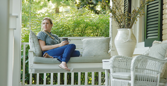 A Caucasian Woman in her Forties Drinks a Cup of Coffee while Sitting on a Porch Swing Outdoors Surrounded by Lush Greenery