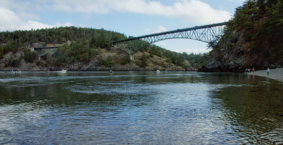 Water Laps the Shore with Deception Pass Bridge Spanning Between Whidbey Island and Fidalgo Island in the Background in Washington on a Partly Sunny Day