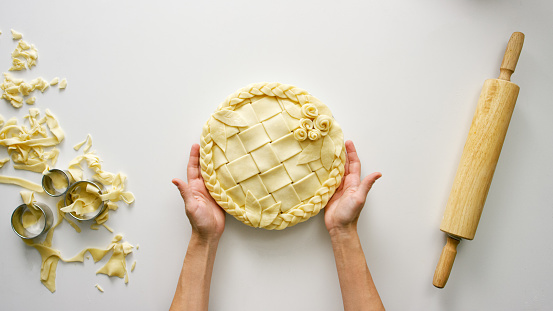 Directly Overhead Shot of a Woman's Hands Holding an Unbaked Lattice Pie with Leaves, a Braid, and Rosettes on a Table Next to a Rolling Pin and Dough Scraps
