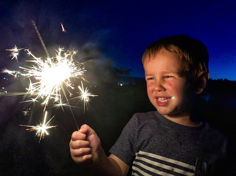 A Cute, Four Year-Old Caucasian Boy Smiles as He Looks at Bright Sparkler on the Fourth of July