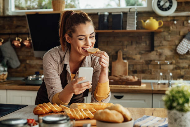istock Happy woman eating slice of bread while using smart phone in the kitchen. 1222085751