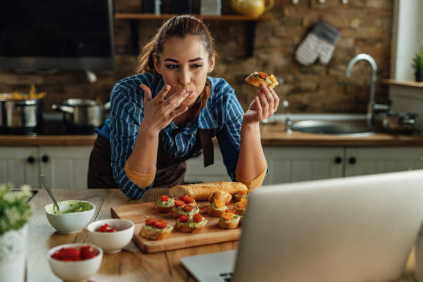 istock Young woman using laptop while eating bruschetta in the kitchen. 1222086392