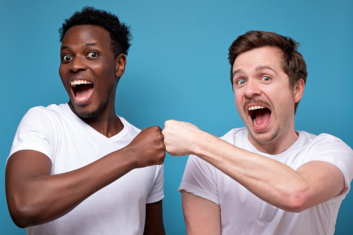 Happy african american and caucasian male friends give fists bump look at camera, smiling make deal show unity and cooperation. Studio shot