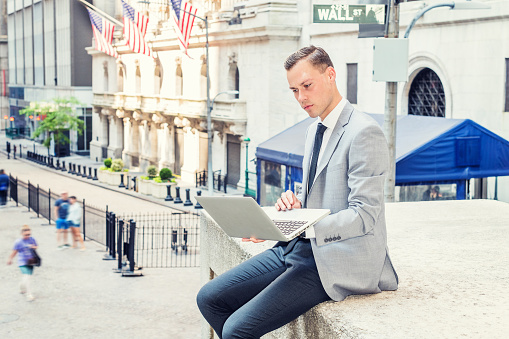 Young Businessman traveling, working in New York City, wearing gray blazer, white shirt, black tie, black pants, sitting on street by Wall Street sign, looking down, working on laptop computer.