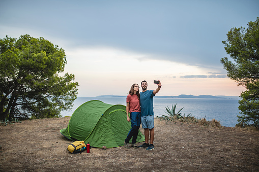 Smiling male and female campers standing next to tent with arms around each other and taking selfie with Mediterranean in background.