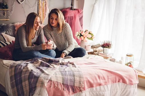 High angle view of young pretty women sitting down on a bed and sharing time together while using a smartphone.