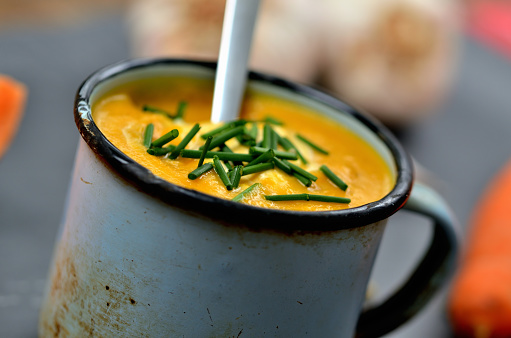 Close-up of pumpkin, carrot soup with cream, fresh chive and spoon in old enamel cup.